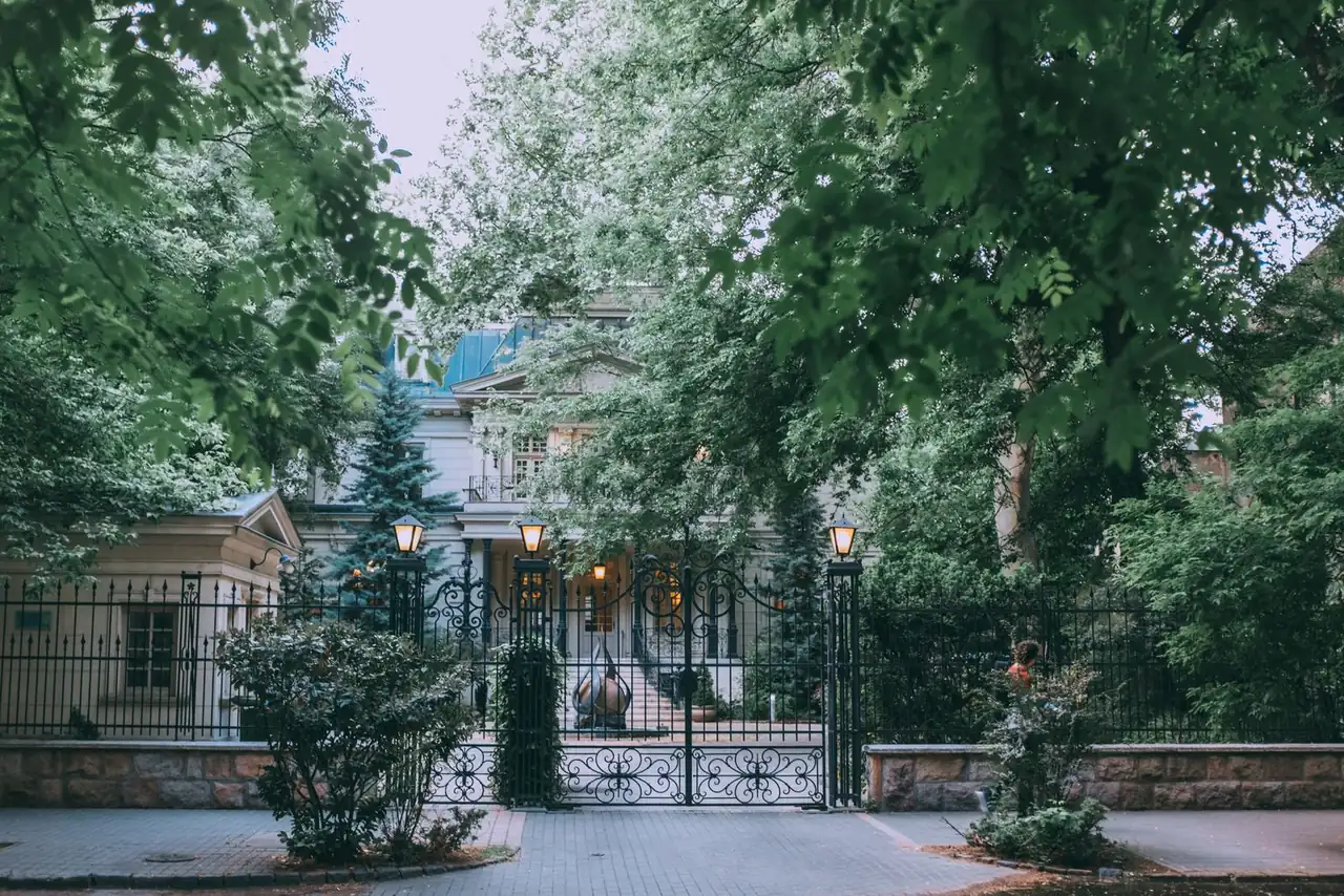 Exterior of stylish classic styled mansion with forged fence and elegant street lamps surrounded by lush green trees in daytime